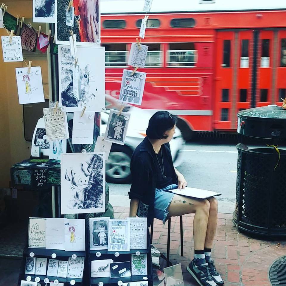 Lex sits at a street-side display of their artworks, as a blurry red trolley bus speeds by